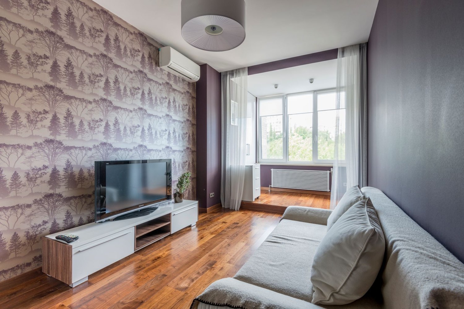 Salon z tapetą w drzewa na ścianie za telewizorem, jasna sofa i drewniana podłoga