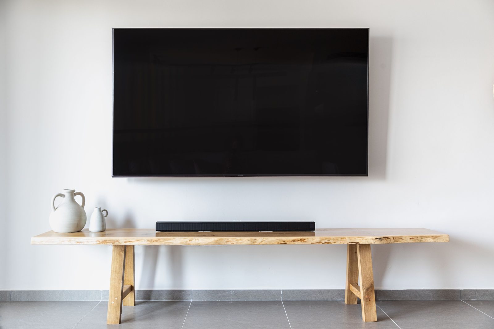 Duży telewizor na białej ścianie nad drewnianą ławką z soundbarem i ceramicznymi dzbankami