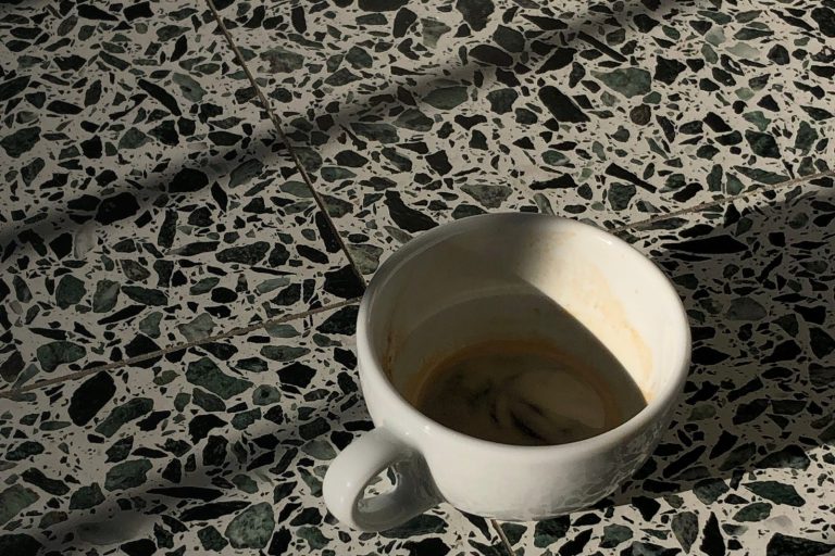 Posadzka z lastryka z dużymi ciemnozielonymi i czarnymi odłamkami kamienia, na niej biała filiżanka w smugach słońca
