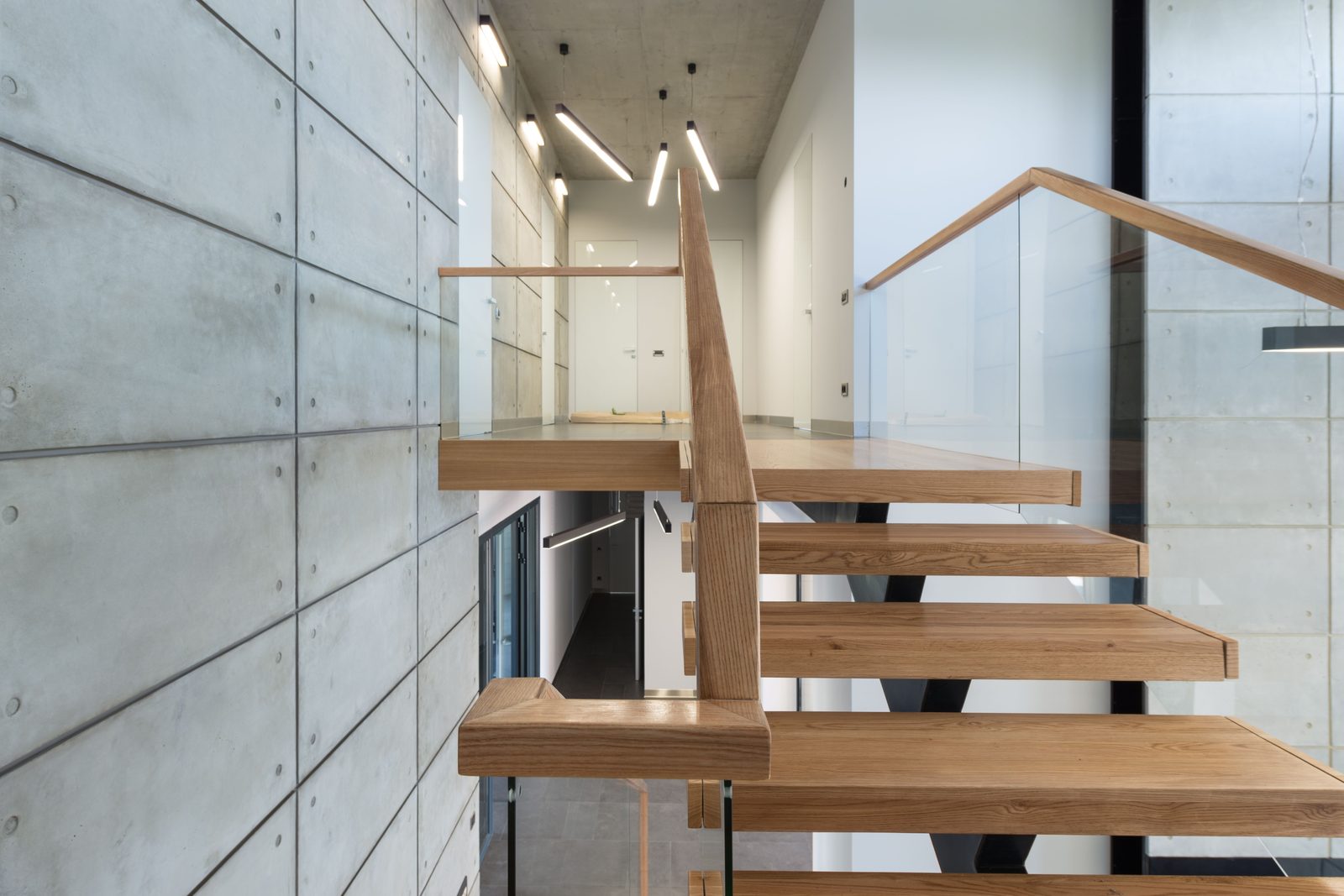 Drewniane stopnie z dębową poręczą i szklaną balustradą przy ścianie z płyt betonowych