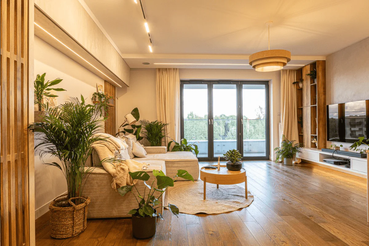 Wnętrze nowoczesnego salonu z roślinami, drewnianymi meblami i dużymi oknami.