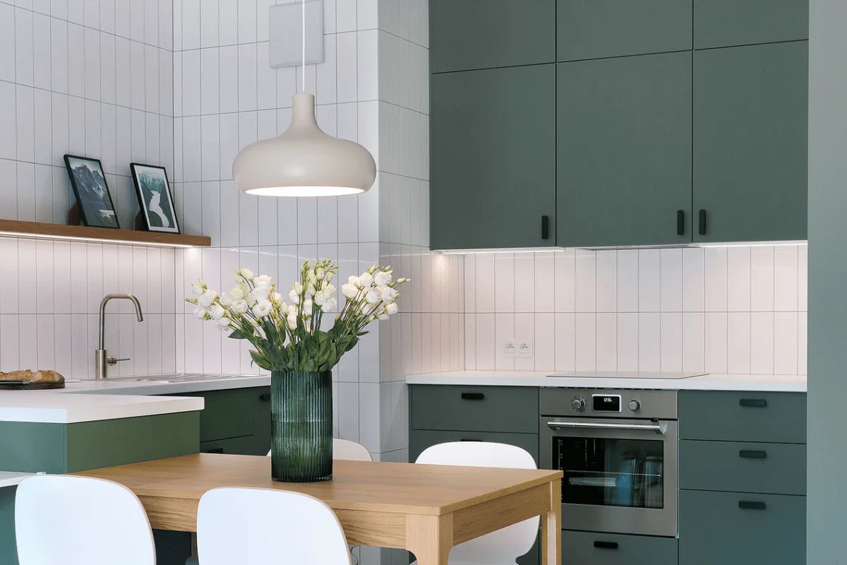 Nowoczesna kuchnia z zielonymi szafkami i drewnianym stołem, kwiaty w wazonie.