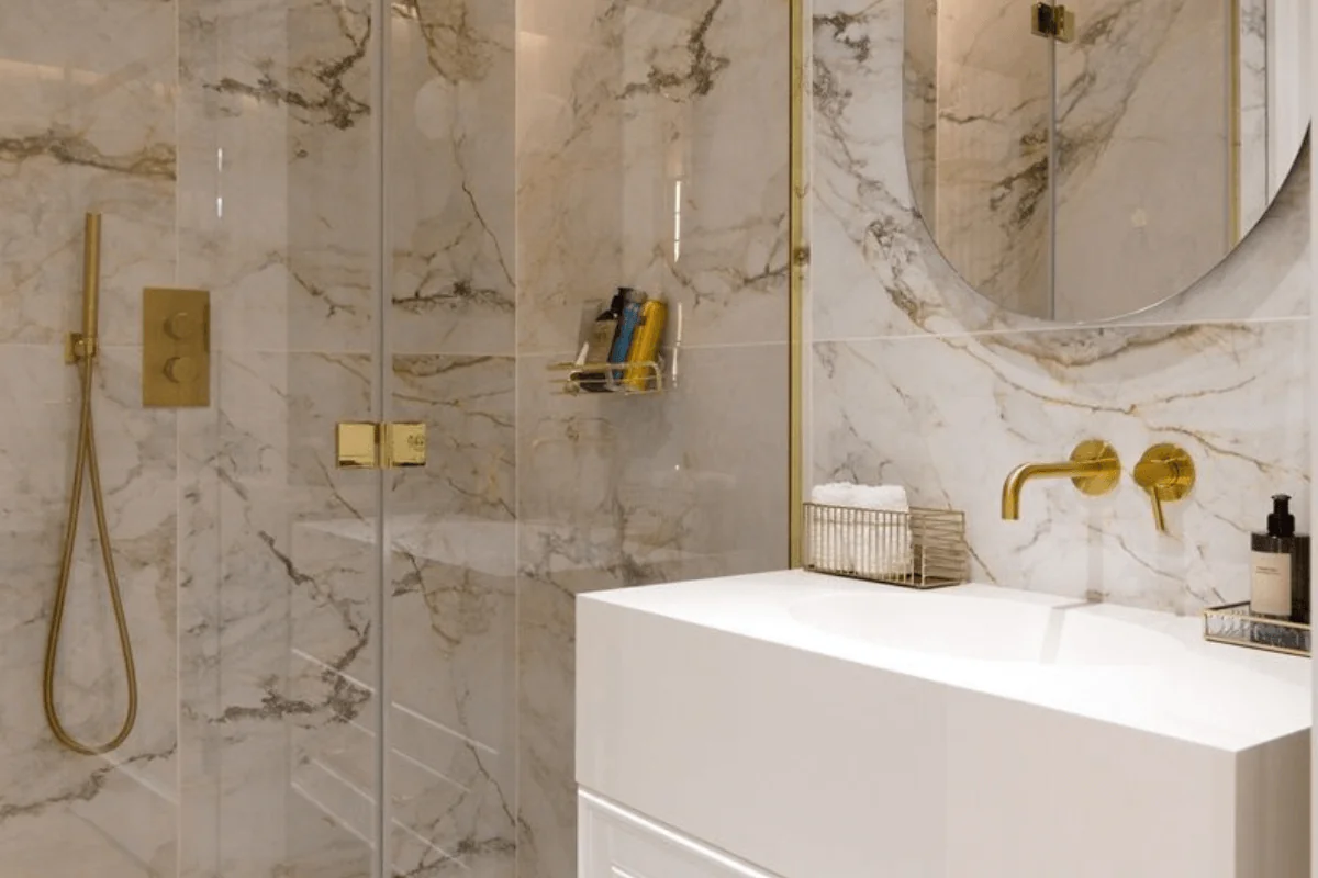 Nowoczesna łazienka z marmurowymi ścianami, złotymi detalami i białą umywalką.