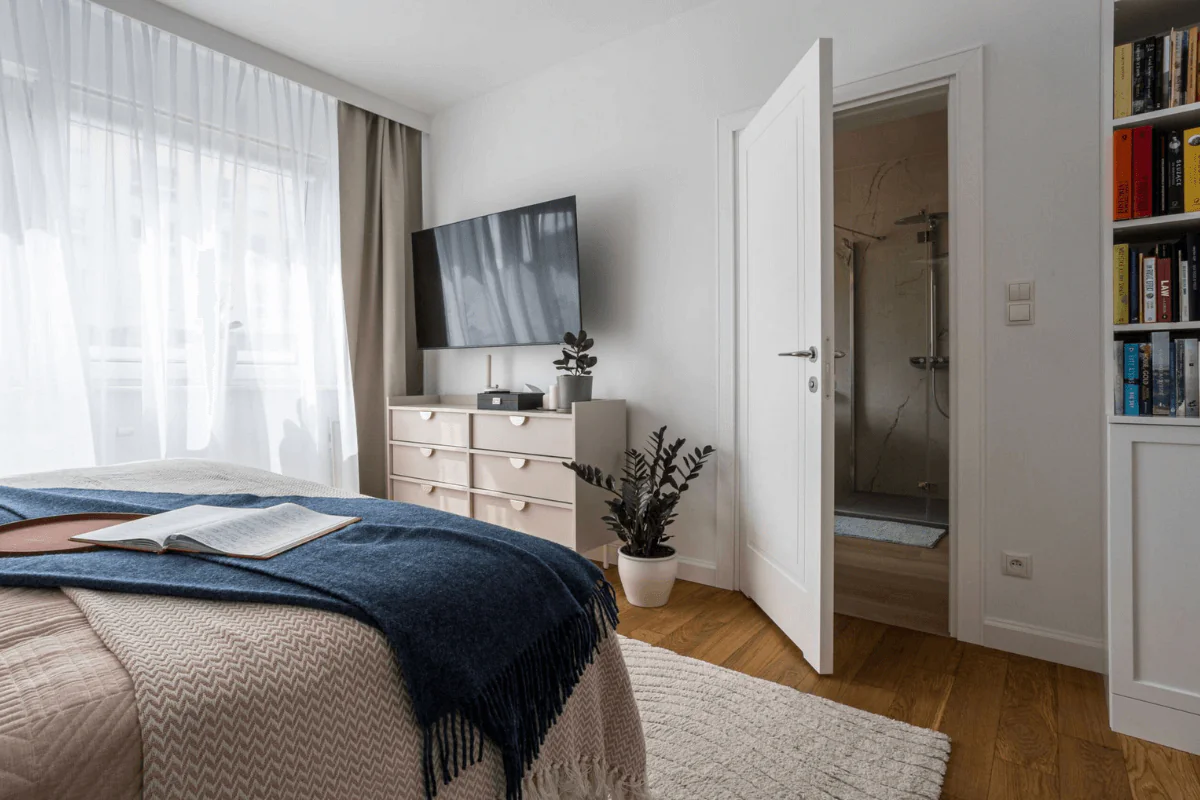 Wnętrze sypialni z białą komodą, telewizorem i zasłonami, styl nowoczesny i tradycyjny.