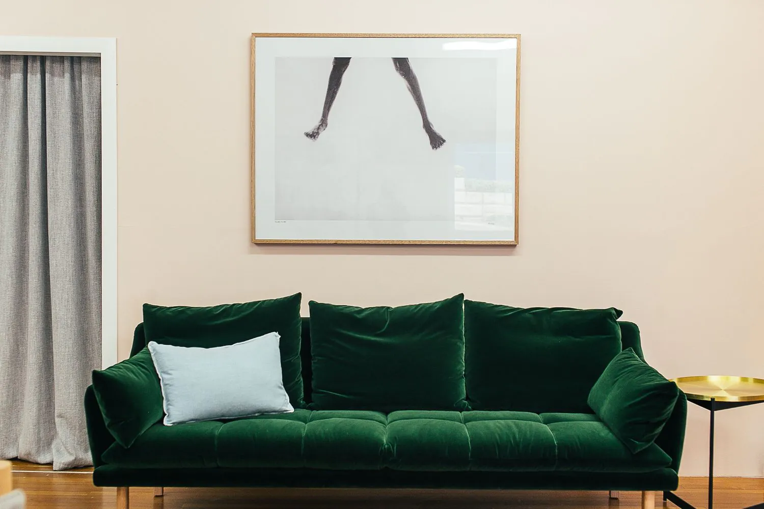 Welurowa sofa w butelkowej zieleni z jasnoniebieską poduszką przy kremowej ścianie z dużą grafiką i złotym stolikiem