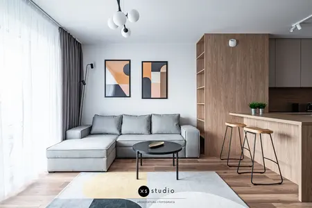 Jasny salon z szarą sofą narożną, drewnianym aneksem kuchennym z hokerami oraz minimalistycznymi dekoracjami na ścianie. Na podłodze geometryczny dywan, a na suficie nowoczesna lampa wisząca.