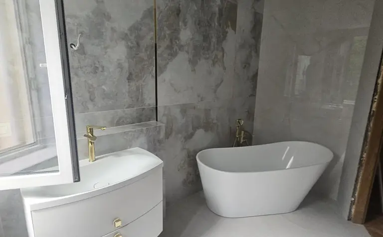 Wnętrze łazienki z białą wanną, szarymi płytkami i złotymi akcentami.