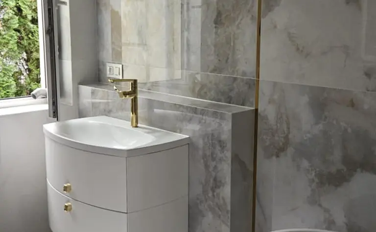 Nowoczesna łazienka z białą umywalką, szarymi płytkami i złotymi akcentami.