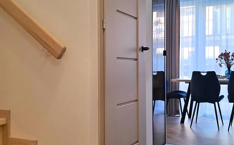 Przedpokój z beżowymi drzwiami, drewnianymi schodami i jadalnią w tle. Styl nowoczesny, skandynawski.