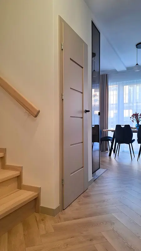 Przedpokój z beżowymi drzwiami, drewnianymi schodami i jadalnią w tle. Styl nowoczesny, skandynawski.