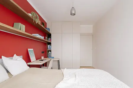 Sypialnia minimalistyczna z czerwoną ścianą, drewnianymi półkami i nowoczesnym biurkiem.