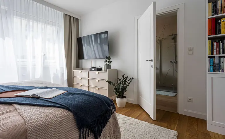 Wnętrze sypialni z białą komodą, telewizorem i zasłonami, styl nowoczesny i tradycyjny.