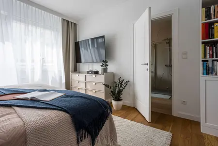 Wnętrze sypialni z białą komodą, telewizorem i zasłonami, styl nowoczesny i tradycyjny.