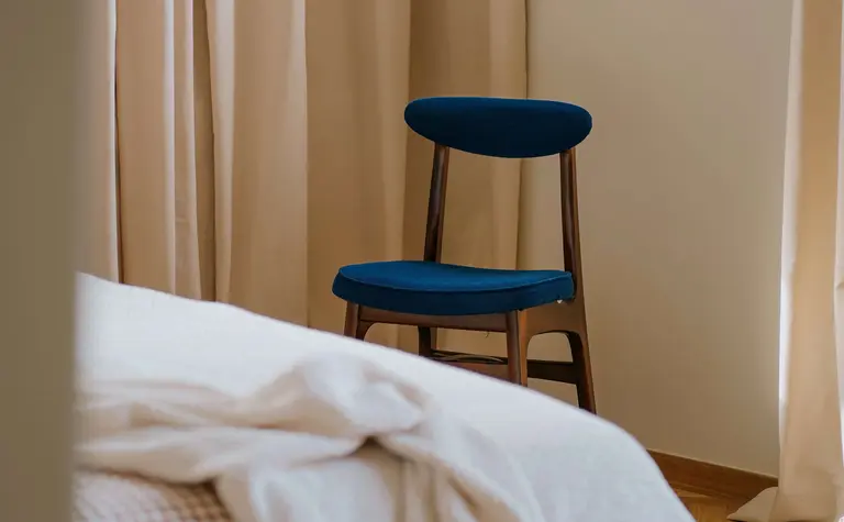 Wnętrze sypialni z beżowymi zasłonami i niebieskim krzesłem w stylu vintage i soft minimalizm.