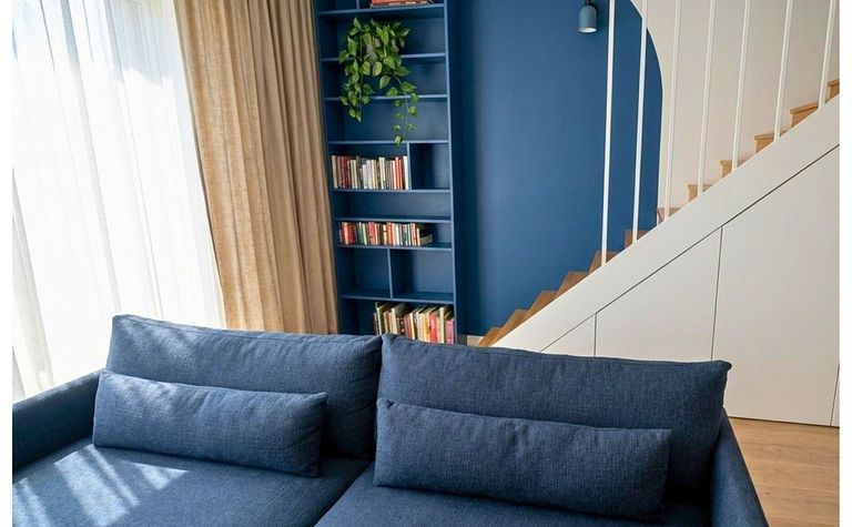 Wnętrze salonu z niebieską sofą, drewnianymi schodami i regałem. Styl boho i eklektyczny.