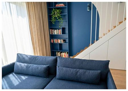 Wnętrze salonu z niebieską sofą, drewnianymi schodami i regałem. Styl boho i eklektyczny.