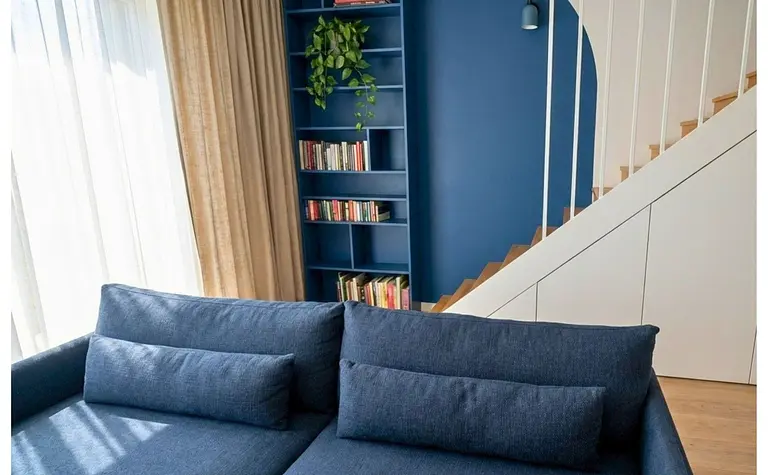 Wnętrze salonu z niebieską sofą, drewnianymi schodami i regałem. Styl boho i eklektyczny.
