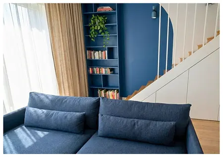 Wnętrze salonu z niebieską sofą, drewnianymi schodami i regałem. Styl boho i eklektyczny.