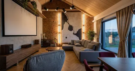 Salon w stylu industrialnym i współczesnym z ceglaną ścianą, szarą sofą, ekranem kina domowego i drewnianą szafką RTV