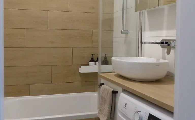 Minimalistyczna łazienka z drewnianymi płytkami, białą wanną i pralką.