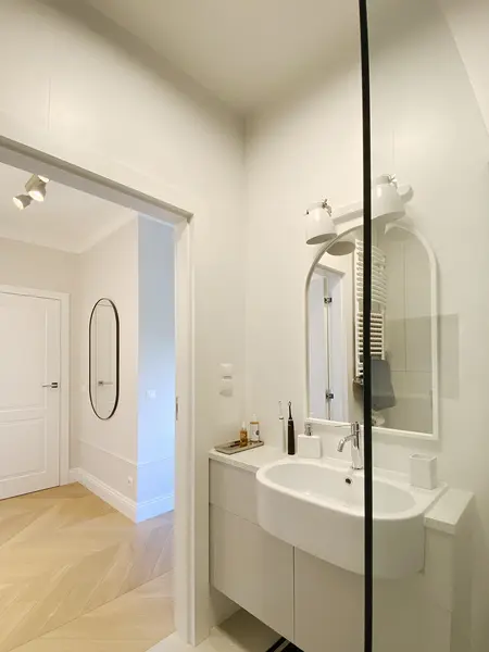 Nowoczesna łazienka z białą umywalką, lustrem i drewnianą podłogą, minimalistyczny design.