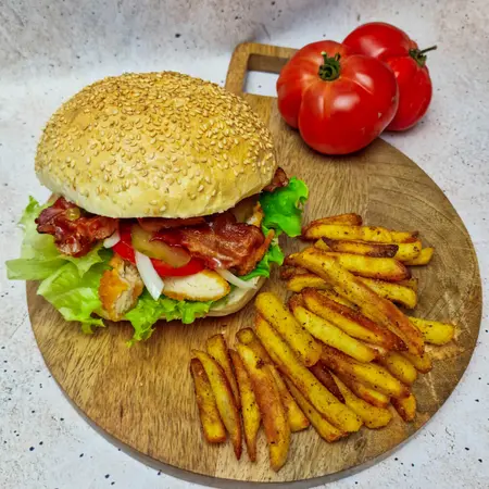 Burger z bekonem, sałatą i pomidorem na desce, obok frytki.