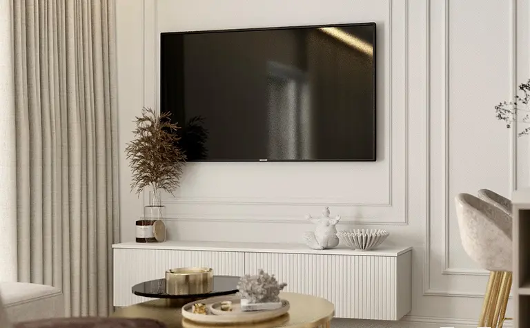 Stylowy salon z telewizorem na ścianie, białymi panelami dekoracyjnymi, złotym stolikiem kawowym i delikatnymi dodatkami w jasnych tonacjach