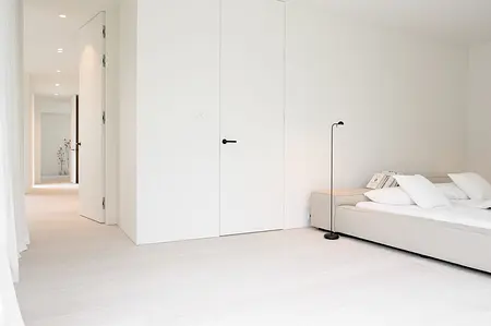 Minimalistyczna sypialnia z białymi ścianami, szarym łóżkiem i nowoczesnym oświetleniem.