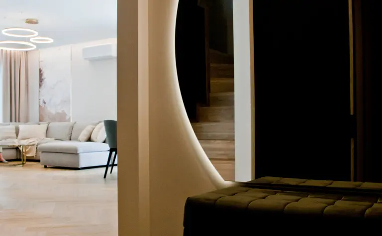 Nowoczesny korytarz z podświetlanym, okrągłym przejściem do salonu, w którym znajduje się jasna sofa i designerskie lampy wiszące. Schody i elegancka ławka w tle dodają wnętrzu stylu.