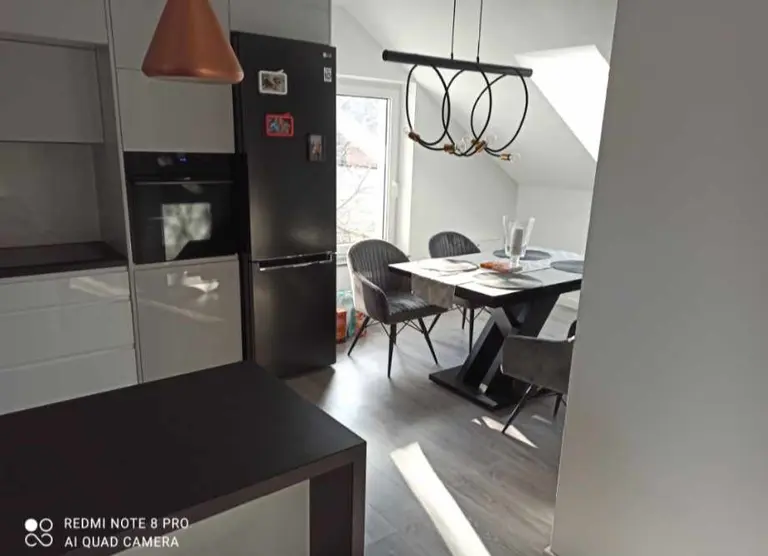 Minimalistyczna kuchnia z czarną lodówką, drewnianym stołem i nowoczesnym oświetleniem.