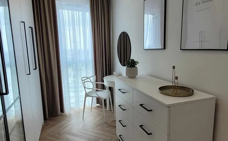 Wnętrze pokoju z białą komodą, okrągłym lustrem i drewnianą podłogą.