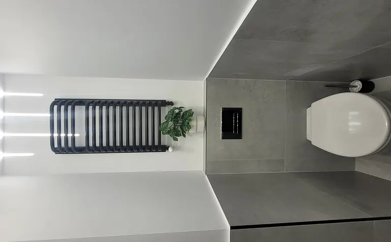 Toaleta z szarymi płytkami, białą toaletą i rośliną w doniczce, minimalistyczny design.