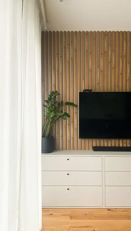 Minimalistyczny salon z drewnianą ścianą, telewizorem i rośliną.