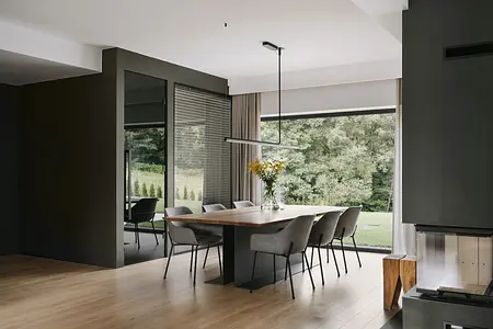 Wnętrze jadalni z drewnianym stołem, szarymi krzesłami i dużymi oknami w stylu industrialnym i minimalistycznym.