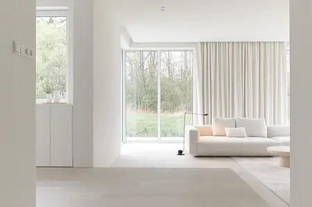 Wnętrze minimalistycznego salonu z białą sofą, dużymi oknami i jasnymi zasłonami.