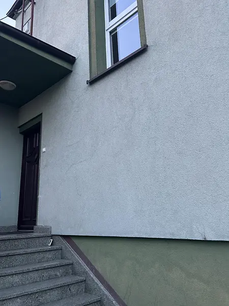Fasada budynku z szarym tynkiem, drewnianymi drzwiami i schodami, styl tradycyjny.