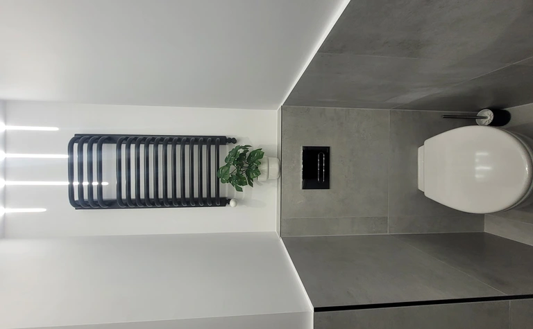Toaleta z szarymi płytkami, białą toaletą i rośliną w doniczce, minimalistyczny design.