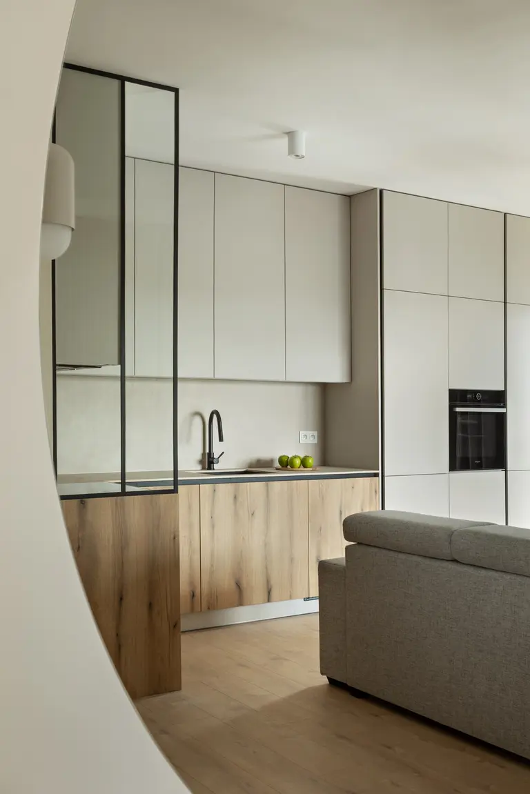 Nowoczesna kuchnia w stylu minimalistycznym z drewnianymi frontami, czarnym kranem i jasnymi szafkami, w tle wbudowany piekarnik. Przestrzeń w stonowanych barwach z eleganckim oświetleniem i akcentami ze szkła.