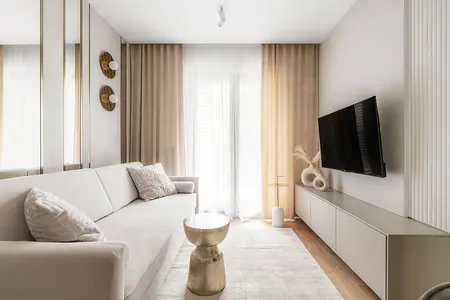 Współczesny salon z beżową sofą, złotym stolikiem i telewizorem na ścianie.