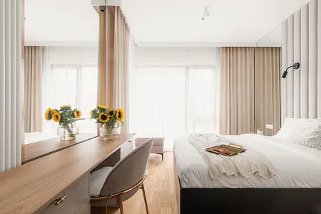 Wnętrze sypialni z jasnymi zasłonami, drewnianymi meblami i kwiatami w wazonie.