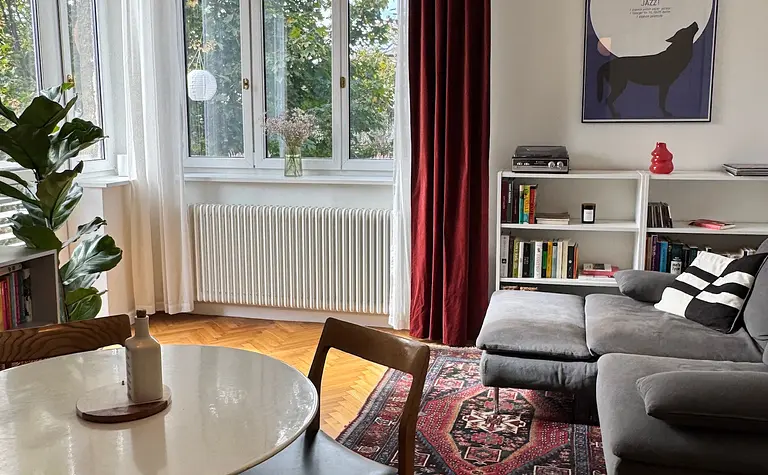 Eklektyczne wnętrze salonu z czerwonymi zasłonami, szarym narożnikiem i drewnianymi akcentami.