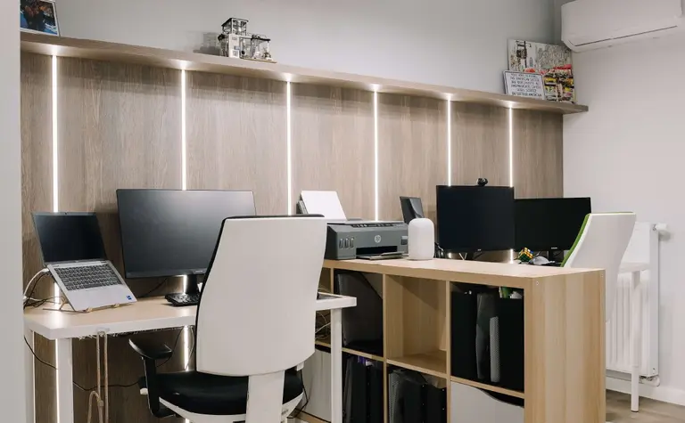 Biuro z jasnymi meblami i nowoczesnym oświetleniem, z komputerami i ergonomicznymi krzesłami.