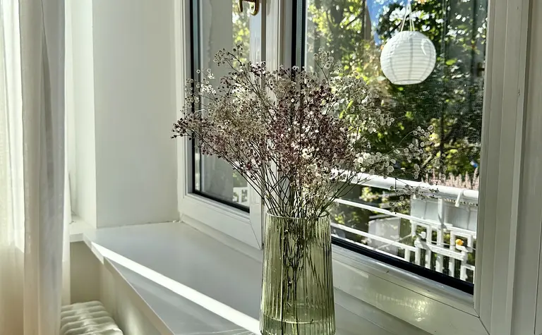 Eklektyczne wnętrze z oknem, zasłonami i suszonymi kwiatami na parapecie.