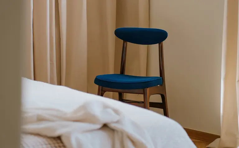 Wnętrze sypialni z beżowymi zasłonami i niebieskim krzesłem w stylu vintage i soft minimalizm.