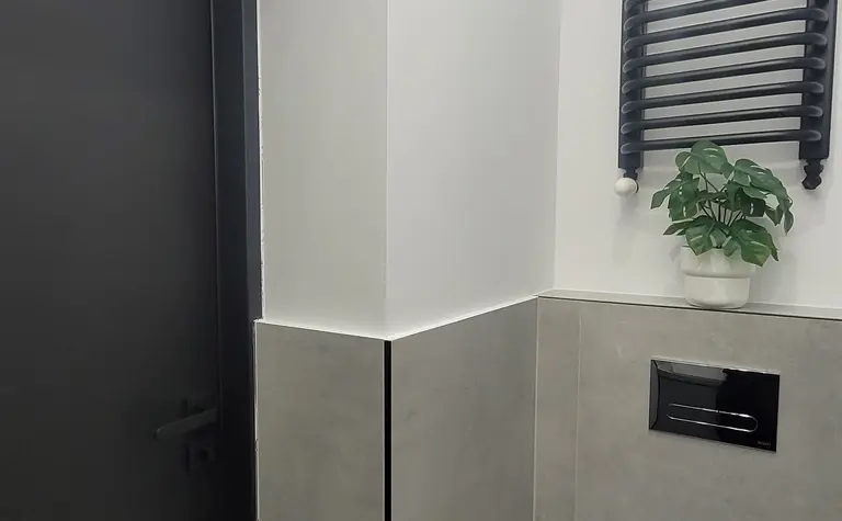 Toaleta w stylu minimalistycznym z szarymi płytkami, czarnymi akcentami i rośliną.