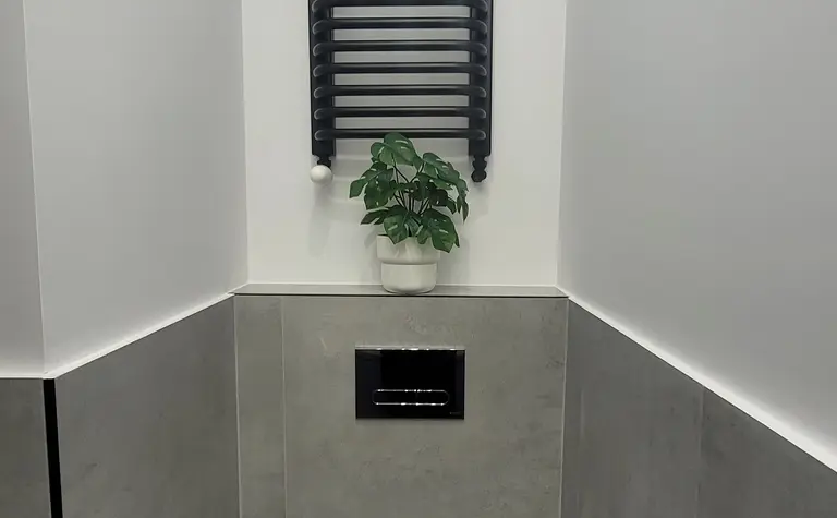 Toaleta z szarymi płytkami, białą toaletą i rośliną w doniczce, minimalistyczny design.