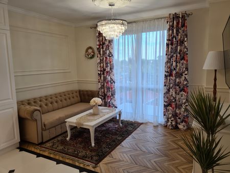 Tradycyjny salon z beżową sofą, kwiatowymi zasłonami i drewnianą podłogą.