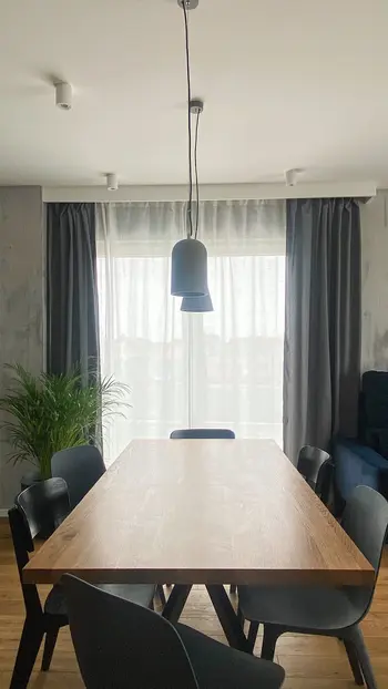Jadalnia w stylu minimalistycznym z drewnianym stołem, szarymi zasłonami i nowoczesnym oświetleniem.