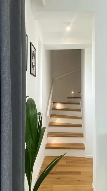Korytarz w stylu minimalistycznym z drewnianymi schodami, roślinami i nowoczesnym oświetleniem.