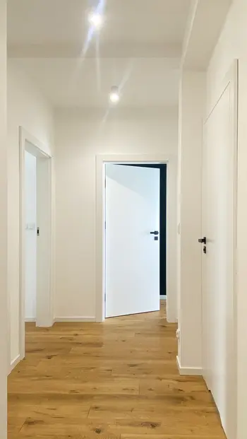 Korytarz w stylu minimalistycznym z białymi ścianami, drewnianą podłogą oraz nowoczesnymi drzwiami.